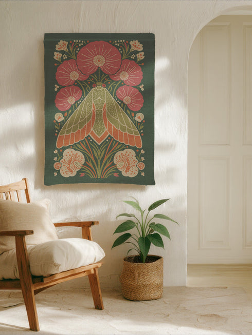 A rectangular woven wall tapestry with a rich, dark green background. The central design is a large, stylized moth with intricate wings in shades of orange, cream, and muted green. The moth is surrounded by a symmetrical arrangement of folk-art style flowers and botanicals in vibrant pink, coral, and cream. The entire piece has a visible, high-quality woven texture. This polyester and cotton blend textile art measures 26x36 inches and includes a rod pocket on the black backing for easy hanging.
